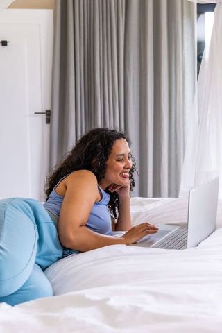 African american woman relaxing on bed using laptop in bright cozy minimalist bedroom