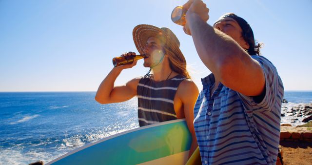 Two young friends are drinking beer by the ocean on a sunny day. One is holding a surfboard while the other is enjoying the view. Ideal for use in summer vacation promotions, lifestyle blogs, or advertisements for beachwear or outdoor activities.