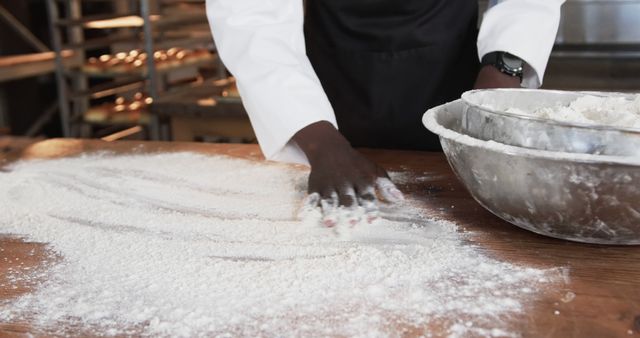 Baker's hands spreading flour on wooden table in bustling industrial kitchen. Ideal for illustrating culinary recipes, demonstrating baking techniques, or highlighting a professional kitchen environment.