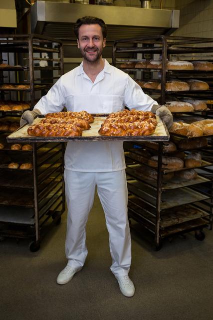 Smiling baker holding loaf of freshly baked twisted buns in bakery kitchen