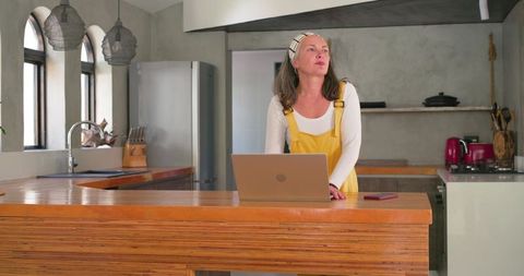 Mature woman in yellow overalls standing at kitchen island using laptop for remote work