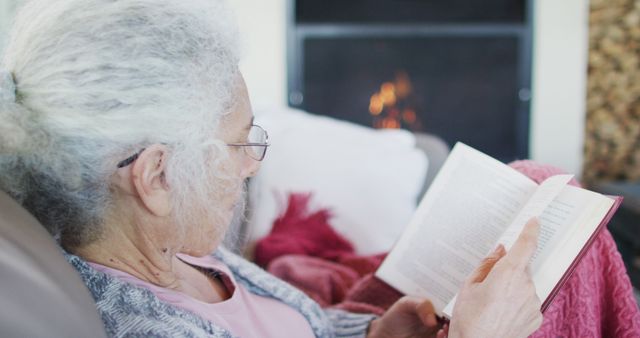 Senior woman is enjoying a quiet evening at home, reading a book next to a warm fireplace. This scene captures a sense of relaxation, tranquility, and warmth, making it perfect for content promoting leisurely activities, aging gracefully, literature, or cozy home environments.