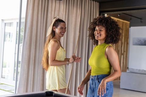 Diverse friends laughing while playing billiards at pool table in fun modern sunlit home