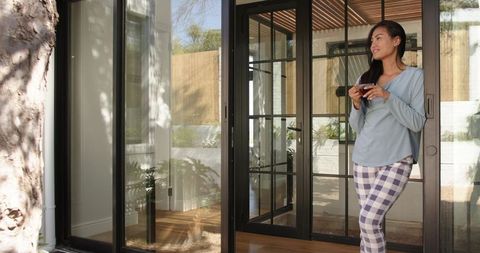 Mid-adult asian woman standing at glass doors in pajamas holding white mug gazing outdoors
