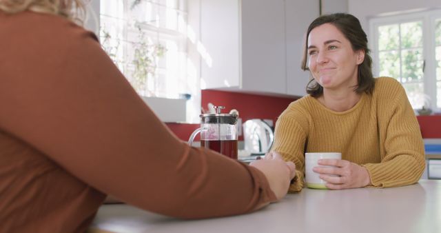 Two women enjoying a conversation at a bright kitchen table with coffee, creating a warm and friendly atmosphere. This image is perfect for lifestyle blogs, social media posts, and promotional materials that highlight relationships, home life, and relaxation.