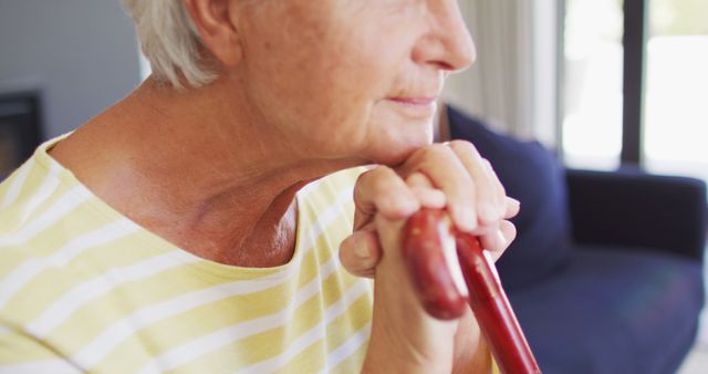 Senior woman sitting and holding a walking stick, staring thoughtfully into the distance. Ideal for topics on elderly care, senior lifestyle, contemplation, aging, and peaceful moments at home.