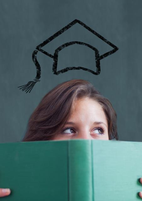 Teenage girl holding book with mortarboard above head against chalkboard