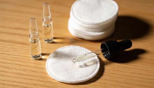 Skincare serum dropper releasing droplet onto cotton pad with ampoules and stacked pads