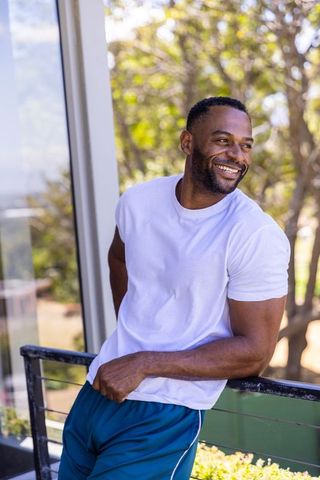 Mature african american man leaning on balcony railing smiling in white tee, teal shorts