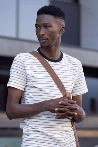 Young african man standing by modern building wearing striped t-shirt and crossbody bag