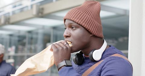 African American Commuter Eating Sandwich Near Office Building Wearing Headphones