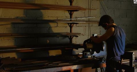 Man cutting long metal stock on bench saw in dim workshop with dramatic shadow and light