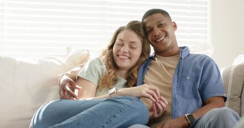 Multiracial couple cuddling on sofa at home wearing smartwatches and casual denim