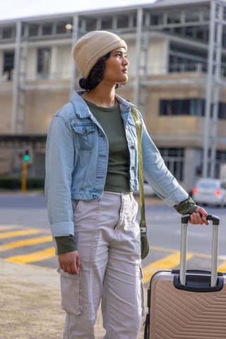 Young urban traveler waiting at crosswalk with suitcase and beanie, city street portrait