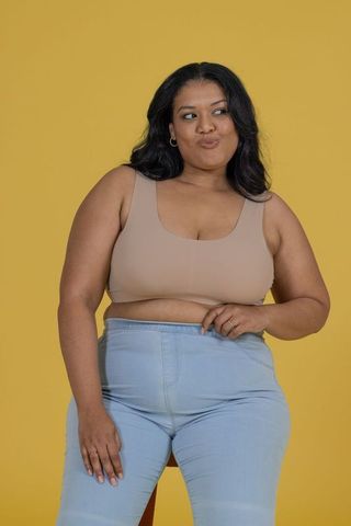 Curvy woman posing on stool in beige crop top and light denim jeans with yellow backdrop