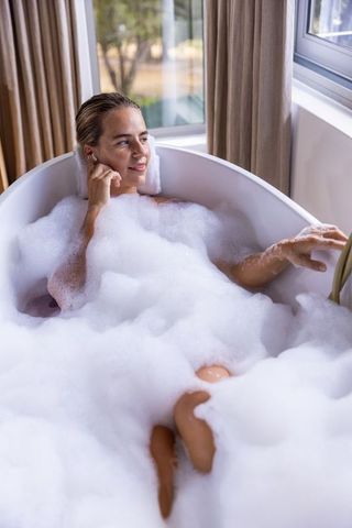 Sunlit bubble bath relaxing woman listening to earbuds in freestanding bathtub