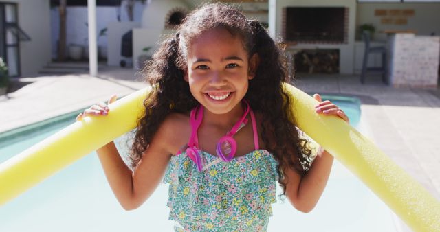 Happy girl with curly hair enjoying summer by the pool. She is smiling and holding a yellow pool noodle, wearing a colorful swimsuit and goggles around her neck. Perfect for advertising summer camps, kid's swimwear, family vacations, or promoting outdoor recreational activities.