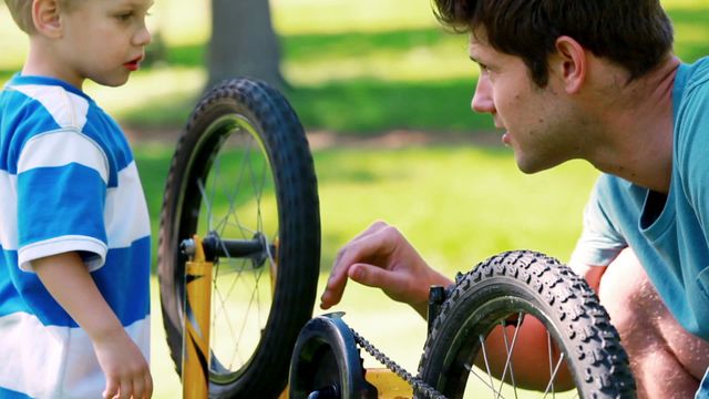 Father and young son bonding through fixing a bike in a sunny park. Video focuses on learning mechanical skills, outdoor activities, and the strong parent-child relationship. Ideal for concepts related to family time, education, parenting, and recreational activities.