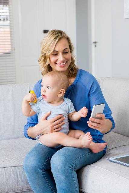 Mother holding baby boy and using mobile phone in living room at home