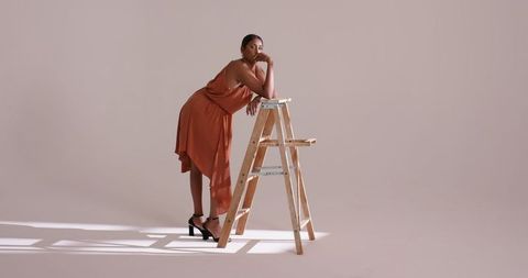 Elegant woman in burnt-orange dress posing on wooden ladder in studio