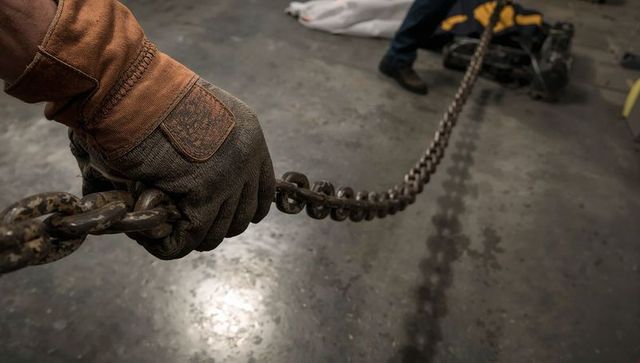 Gloved hand pulling rusty heavy chain across workshop floor with winch and work boots