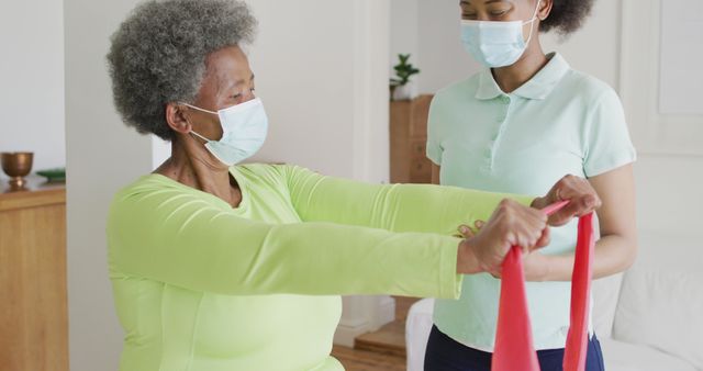 Senior woman wearing mask exercising with resistance band, guided by female physical therapist in home setting. Ideal for depicting home healthcare, senior rehabilitation, and safe indoor fitness routines during pandemic.