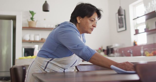 Senior biracial woman wearing apron and cleaning table in kitchen alone - Download Free Stock Photos Pikwizard.com
