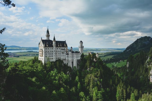 This stock photo features Neuschwanstein Castle perched high above a lush green forest with scenic hills in the background. The castle's towers and turrets stand tall against a partly cloudy sky. Ideal for use in travel blogs, historical articles, brochures, travel guides, and promotional materials related to European tourism, heritage sites, or architectural beauty.