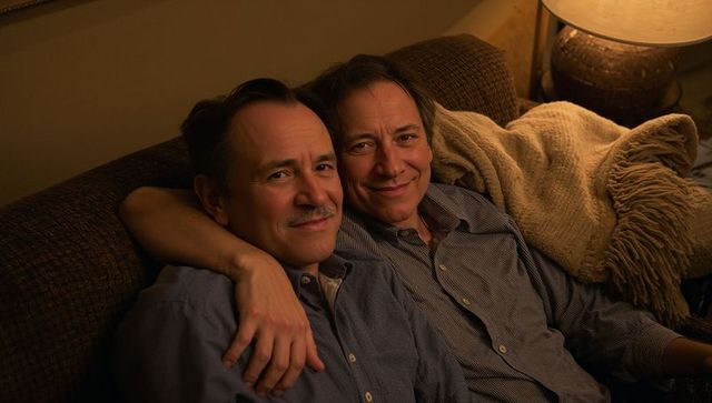 Middle-aged same-sex couple cuddling on couch, smiling and relaxing during cozy warm evening