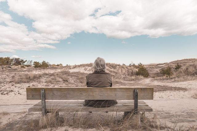 Elderly woman sits alone on a wooden bench overlooking grassy dunes under a partly cloudy sky. Useful for themes of solitude, reflection, nature appreciation, retirement, and tranquility.