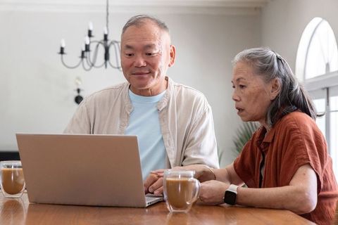 Senior couple collaborating on laptop over coffee at wooden dining table, home lifestyle