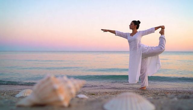 Woman practicing yoga dancer pose on serene sunrise beach with seashells and flowing white outfit