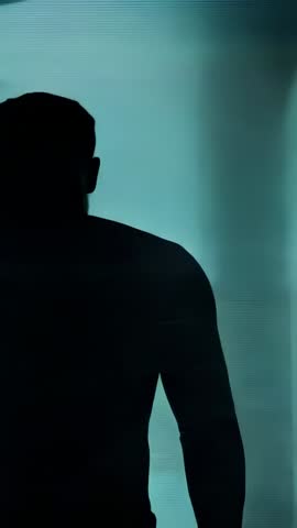 Vertical video showing backlit silhouette man standing and turning head against glowing teal panel f