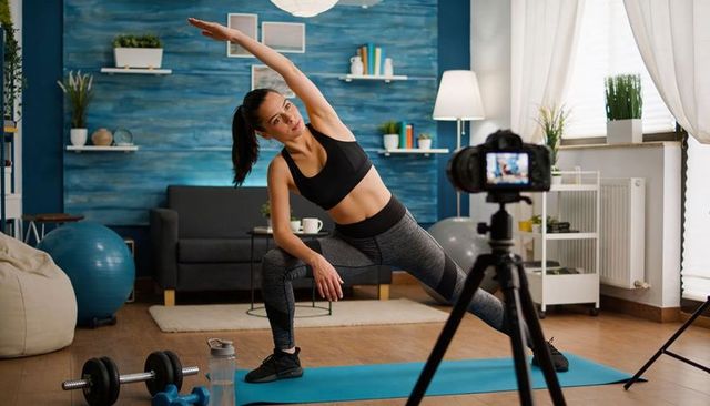 Home fitness trainer stretching while filming workout on yoga mat in living room