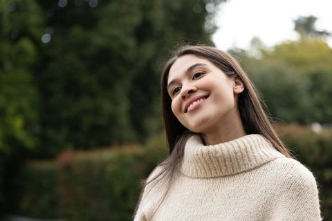 Smiling young woman tilting head in cream knit turtleneck sweater outdoors park portrait