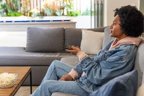 African american woman relaxing on sofa watching tv with remote and bowl of popcorn