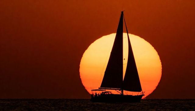 Sailboat silhouette sailing at sunset with oversized sun on horizon