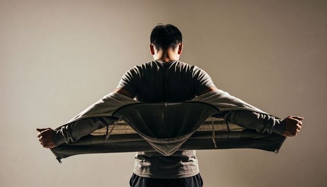 Asian man pulling off grey hoodie revealing dark tee back view rimlight minimal studio