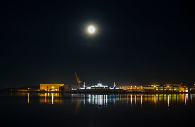 Moonlit waterfront harbor with illuminated buildings at night. Moon glows brightly above, reflecting in calm water, showcasing the architectural beauty of the harbor and its surroundings. Ideal for use in travel magazines, cityscape features, or maritime promotions highlighting nightlife.