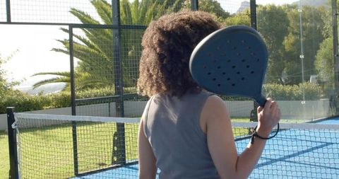 African american woman holding padel paddle on blue court, back view, outdoor athlete