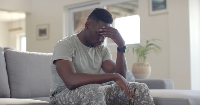 An afro-american military veteran sitting on a couch, looking stressed and lost in thought. He is holding his forehead with one hand, wearing army camouflage pants and a neutral-colored t-shirt. Use this image for articles, blogs, or campaigns about veteran mental health issues, PTSD, support for soldiers, or loneliness.
