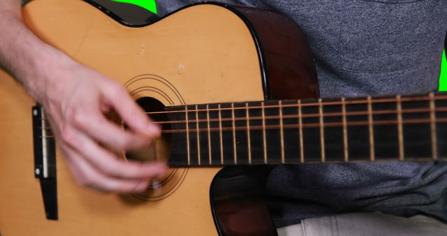 Close-up shows hands strumming acoustic guitar with green screen serving as background. Ideal for music tutorials, online music lessons, promotional videos about musical instruments, and videos requiring a customizable background.