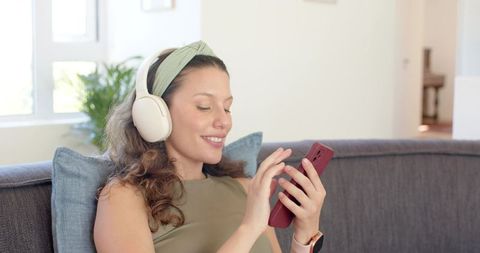 Young woman relaxing on sofa listening with wireless headphones while using smartphone