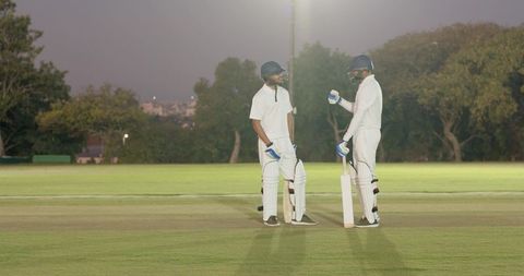 Two batsmen discussing strategy on cricket pitch at dusk under floodlights