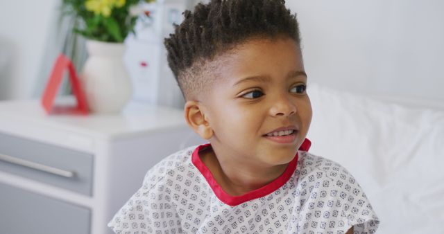 African American boy sitting on a hospital bed, wearing a patterned white and red hospital gown, and smiling. Background includes bedside table with flowers and blurred medical equipment. Ideal for use in healthcare, pediatric care, child well-being, and medical services adverts or brochures.
