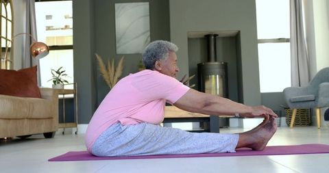 Senior woman stretching forward on yoga mat in sunlit modern living room with wood stove