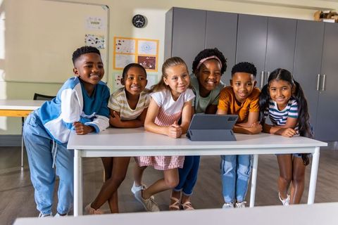 Multicultural kids gathering around tablet in classroom smiling and learning together