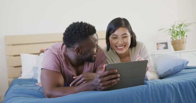 Happy diverse couple using tablet and lying in bedroom. Spending quality time at home concept.