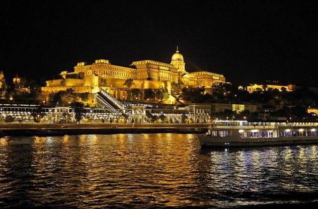 Budapest Castle Hill illuminated by lights at night, reflecting in the Danube River with a boat passing by. This image emphasizes the historical and architectural beauty of Budapest, perfect for use in travel brochures, tour promotions, and culture-focused advertisements highlighting the charm and allure of Hungary's capital city at night.