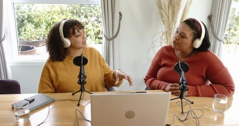 Co-hosts recording podcast at home table, wearing headphones, using laptop and microphones